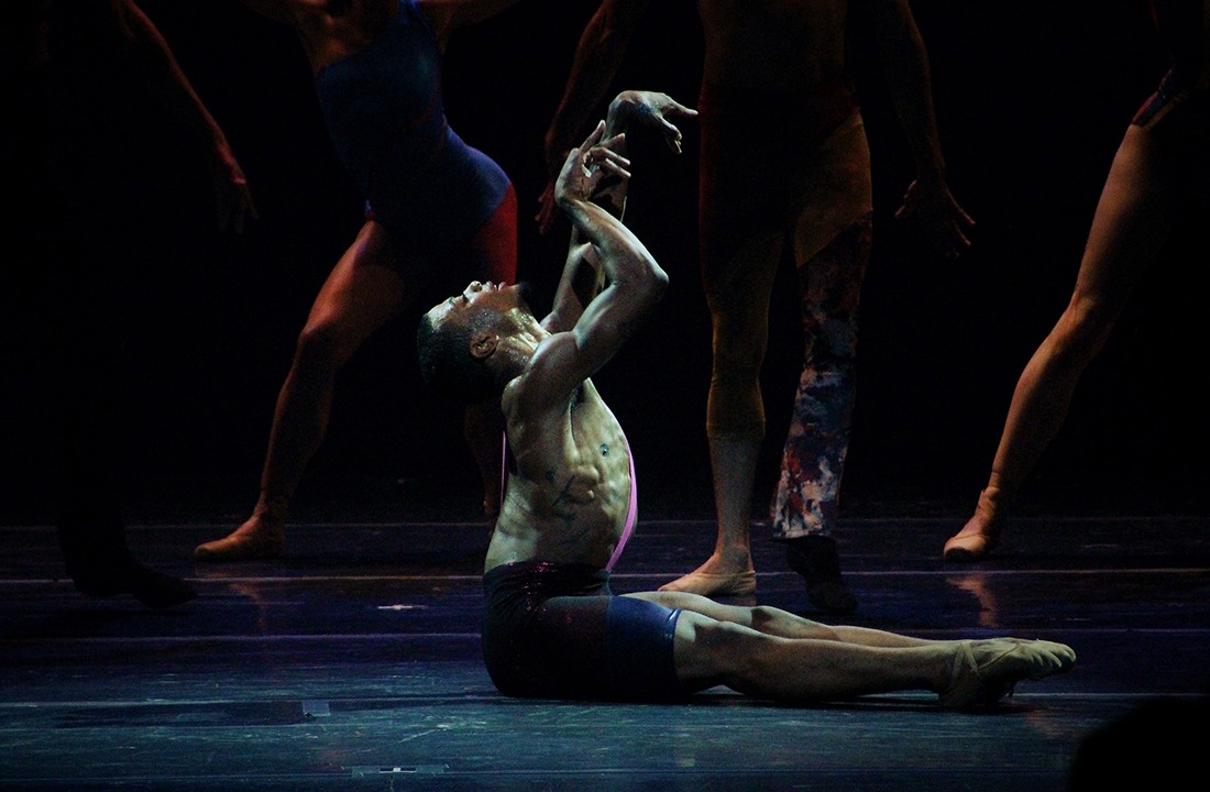 Image of a male performer with no shirt and black shorts seated on the stage with hands raised looking toward the sky while the legs of other performers are visible in the background.