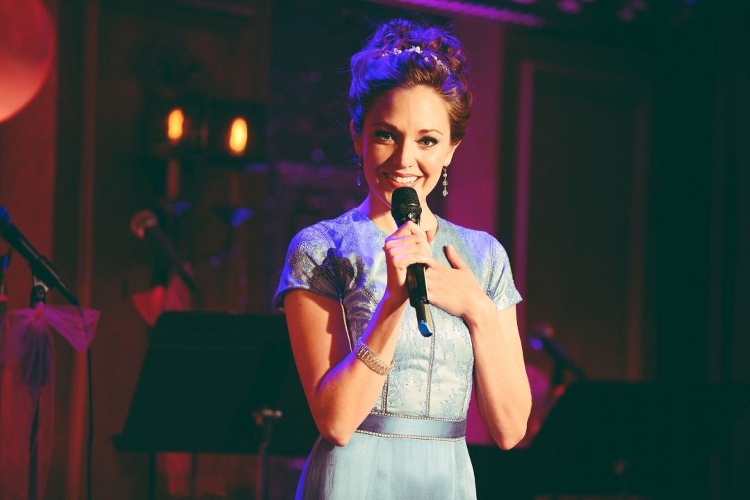 A woman in a light blue dress with a microphone smiling into the camera