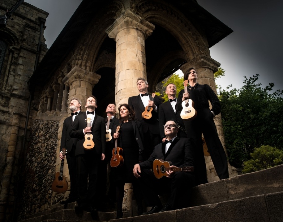 Image from a point of view looking up at the eight ukulele players who are standing outside on steps and holding their ukuleles while looking in various directions with pillars in the background