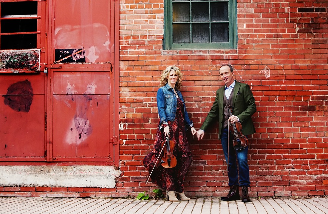 Image of the two performers from Celtic Celebration holding hands and their fiddles while standing in front of a brick wall.