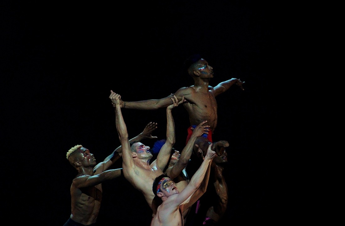 Image of one male performer without a shirt being held aloft by five other shirtless performers