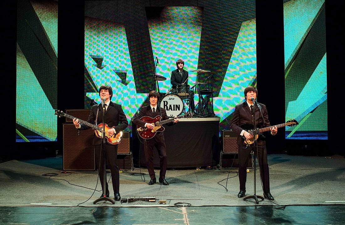 Image of the performers in RAIN on stage in black suits, white, shirts, and black ties in tribute to the early Beatles.