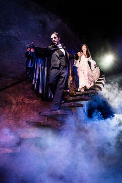 Phantom in white mask and black suite holding a lantern and leading Christine, who is wearing a white dress, down stone steps with a dark background and fog beneath them.