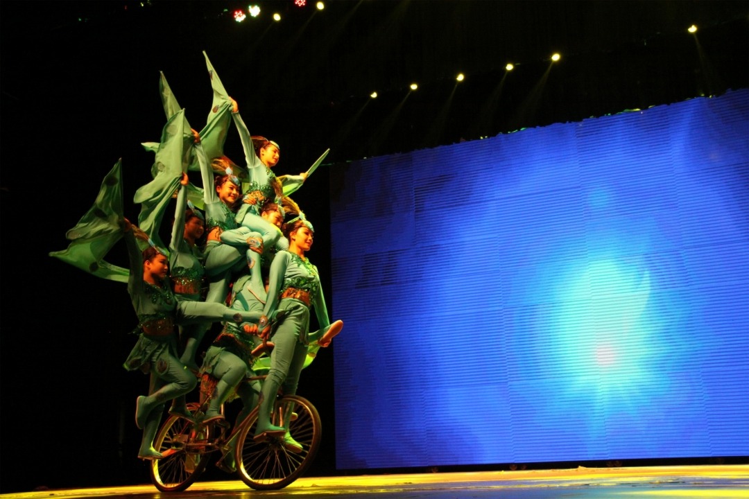 Eight acrobats wearing green costumes riding a single bicycle with a blue light background.