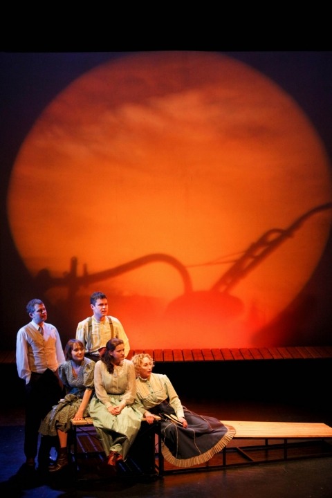 Five people sit and look out as if they are watching what is happening on the screen behind them, which is an orange-tinted image of a landscape with a plow