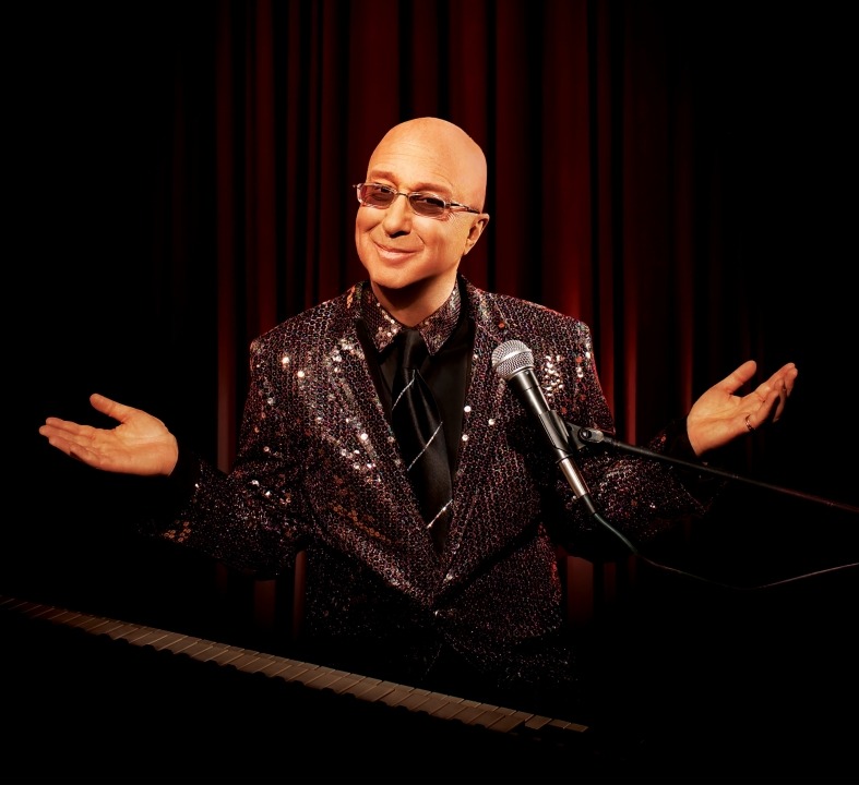 Paul Shaffer posed behind his keyboard and microphone wearing a sequin suit and shrugging to the camera