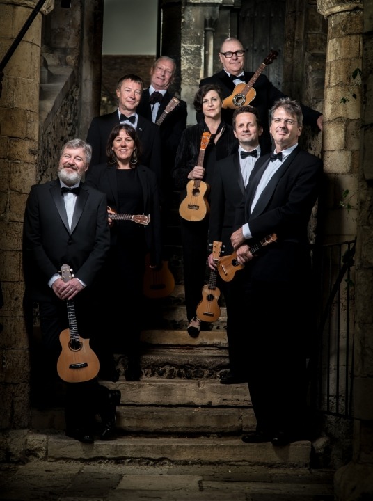 Image of the eight ukulele players holding their ukuleles and posed on steps smiling into the camera with stone walls behind them
