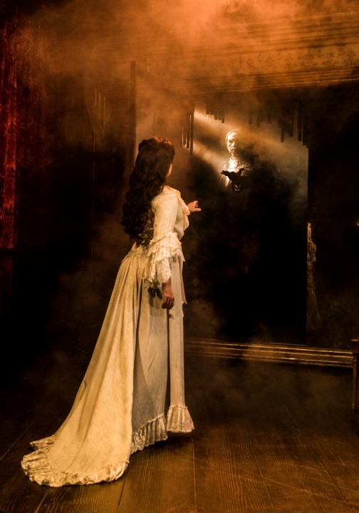 Christine in a white dress facing a mirror that shows the Phantom in black reaching out to her with fog in between them.