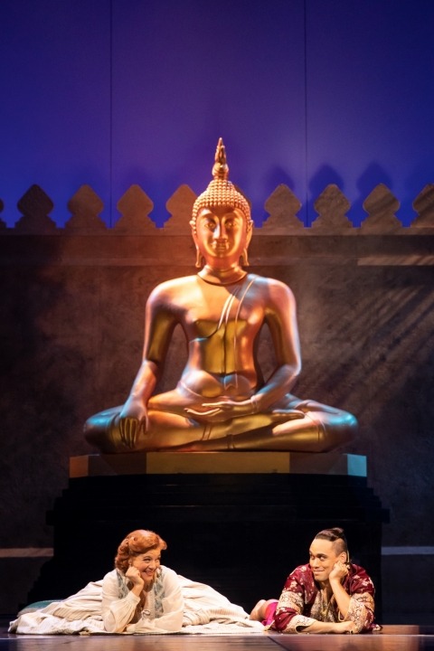 Woman in a white dress on the left and a man in red looking at each other with a statue in the background.