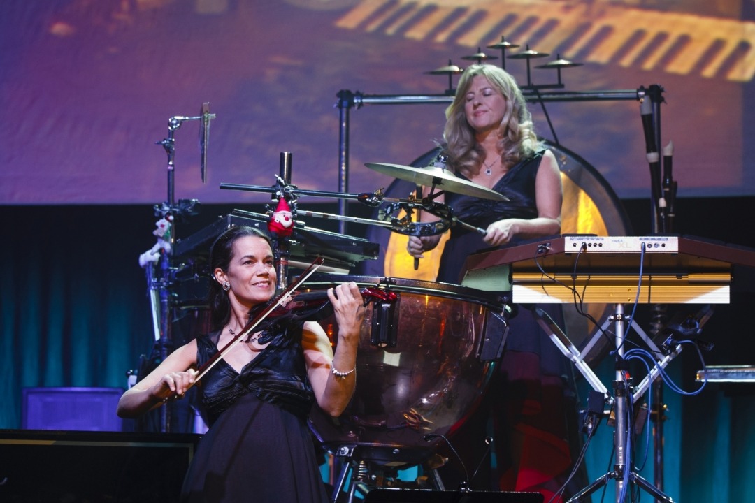 Woman playing a violin and another woman drumming behind her