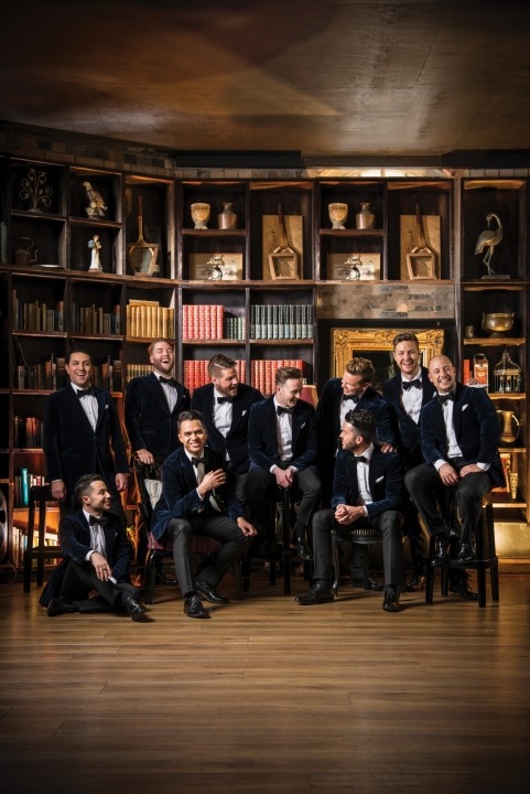 Ten men in blue velvet suits and bow ties sitting with each other and laughing with a grand bookcase in the background