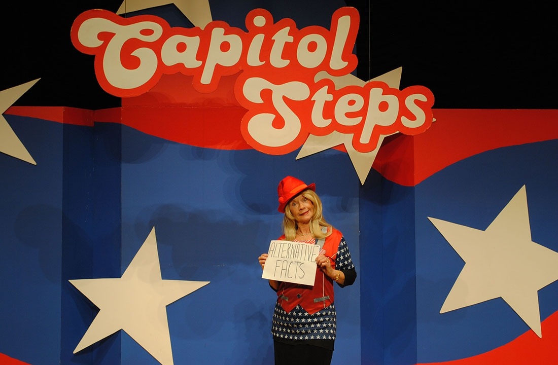 Actor dressed up as Kellyanne Conway wearing a bright red hat and holding a sign that says "Alternative Facts." She is standing in front of a red, white and blue background with Capitol Steps displayed overhead