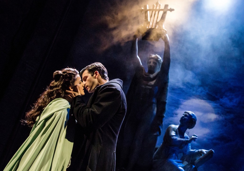 Christine, wearing a green cloak, and Raul, wearing a black coat almost kissing in front of a foggy, dark background with statues behind them.