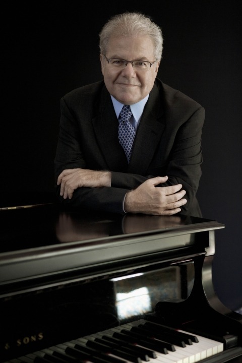 Emanuel Ax stands behind his grand piano and leans forward on it while he smiles at the camera