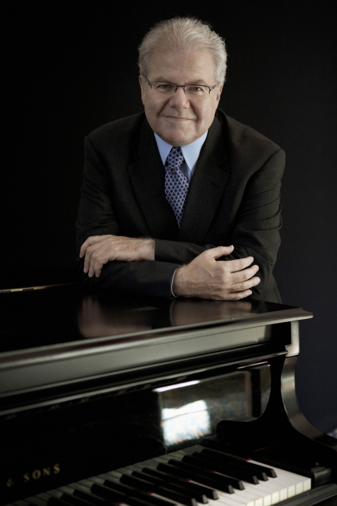 Emanuel Ax stands behind his grand piano and leans forward on it while he smiles at the camera