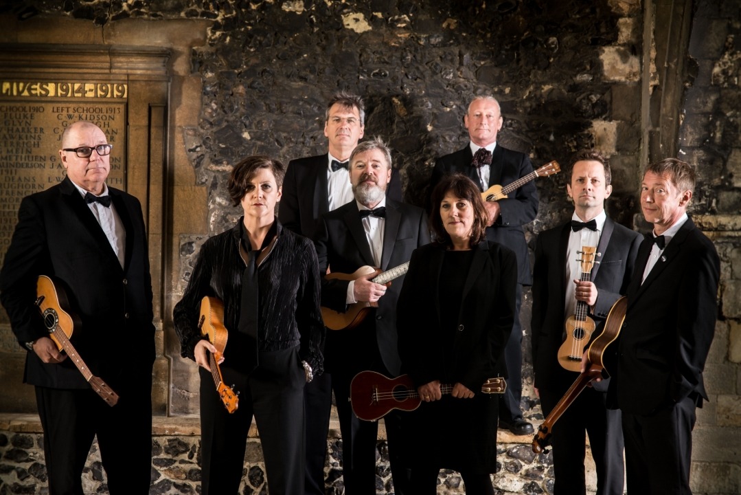 Image of the eight ukulele players holding their ukuleles standing and looking into the camera with exposed brick in the background