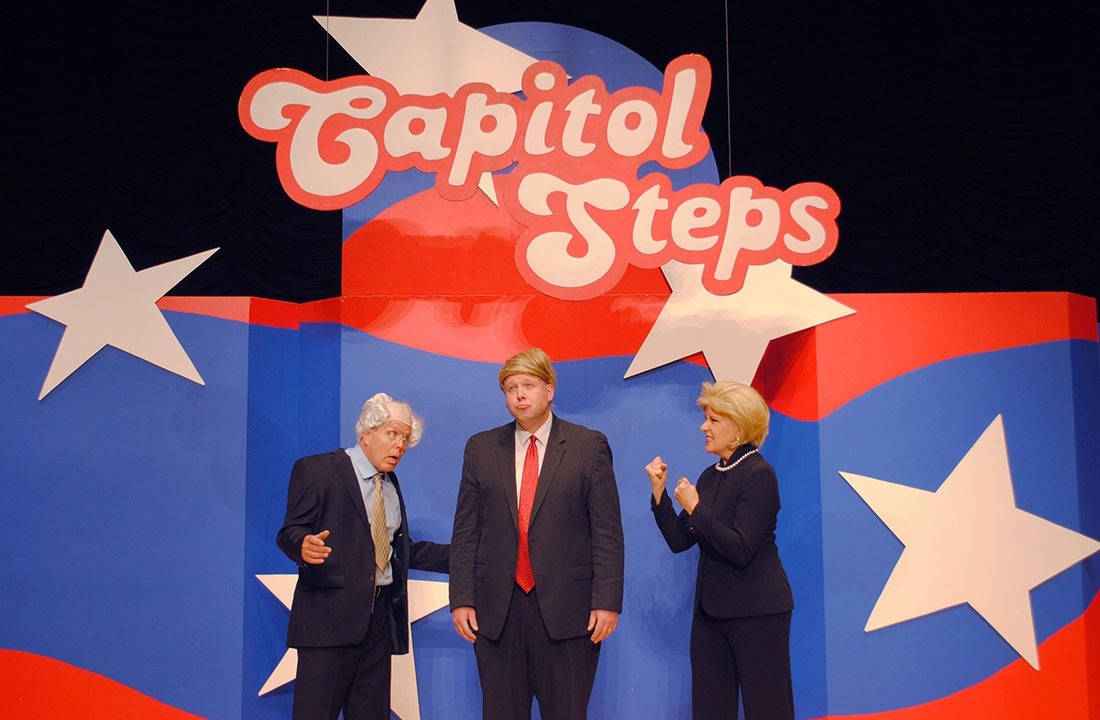 Two actors dressed up as Bernie Sanders and Hillary clinton acting upset at a third actor dressed as Donald Trump; all standing in front of red, white and blue background with Capitol Steps displayed overhead.