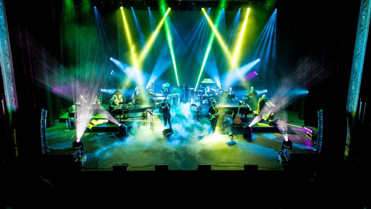Band members performing on a stage with vibrant green, yellow and blue stage lights and fog on the floor