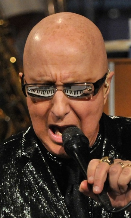 Close up of Paul Shaffer wearing a black sequin suit and singing into a microphone with his keyboard reflecting in his glasses