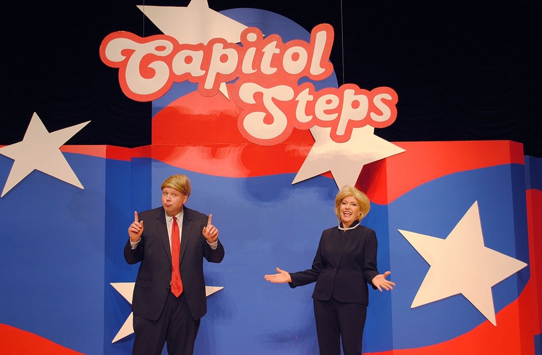 Two actors dressed as Donald Trump and Hillary Clinton debating one another in front of red white and blue background with Capitol Steps displayed overhead
