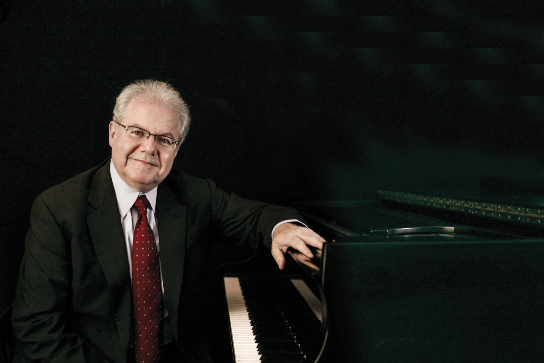 Emanuel Ax in a black suit sitting at his piano with a black background