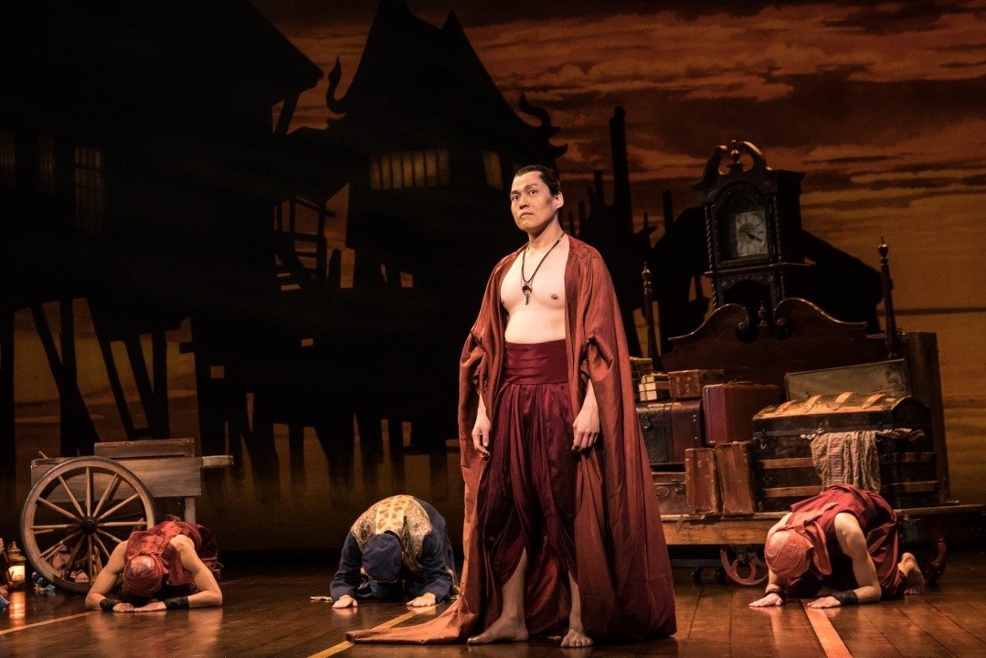 Man in costume singing with two people bowing behind him and a brown/orange background.