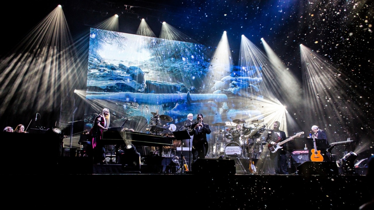Band members playing on stage while white stage lights shine down on them and fake snow falls down. A screen behind the band projects a frozen landscape