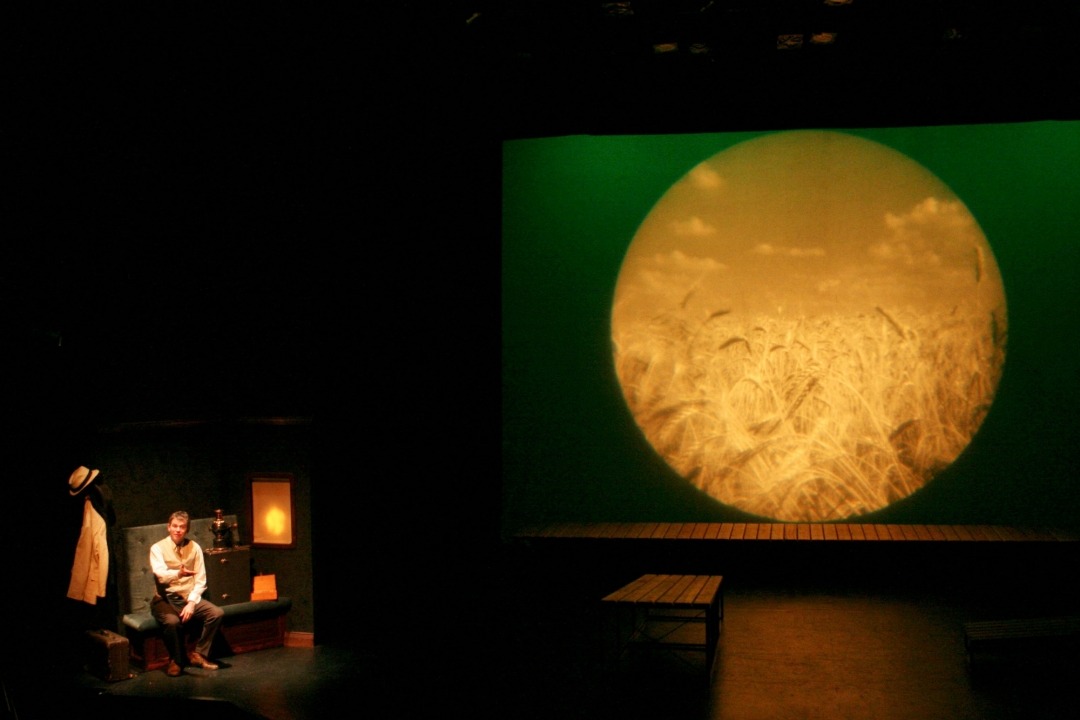 A man sitting in a room to the left of the screen. The screen projects an image of wheat