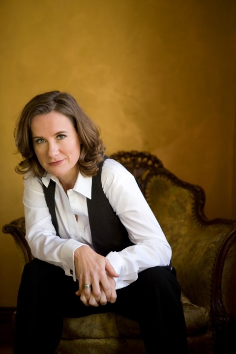 Susan Werner in a white button up and black vest and pants sitting in a luxe chair with a mustard background