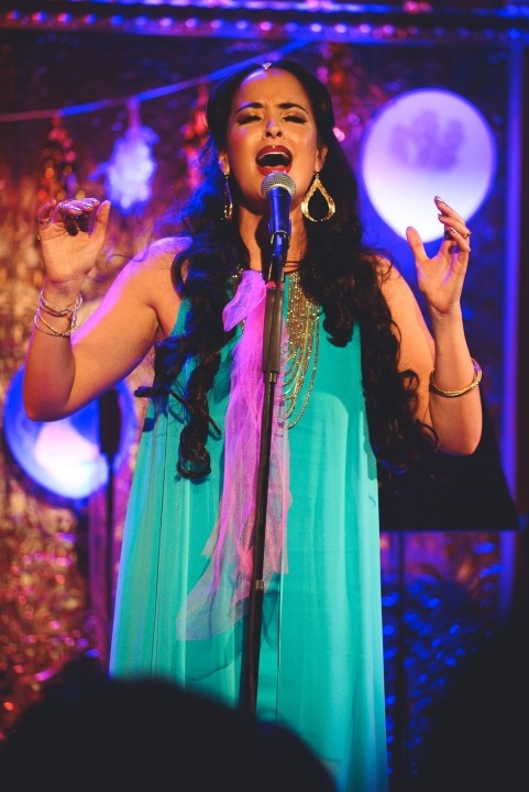 Woman in a turquoise dress and gold jewelry sings into a microphone with a glittery background