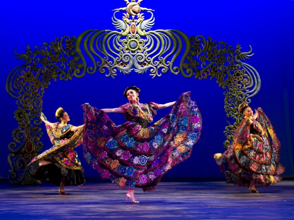Three woman in colorful Mexican attire dance with a flora and fauna style arch in the background