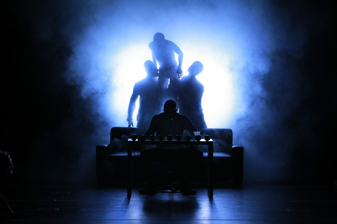 A black silhouette of a many sitting at a desk with three black silhouettes behind him in front of a black and blue smokey background.