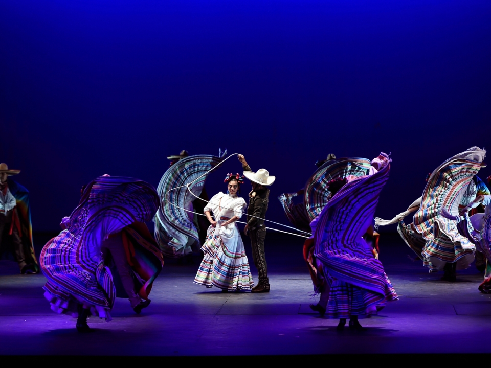 A man and woman in traditional Mexican costume perform a dance with a lasso in a spotlight as other couples dance around them
