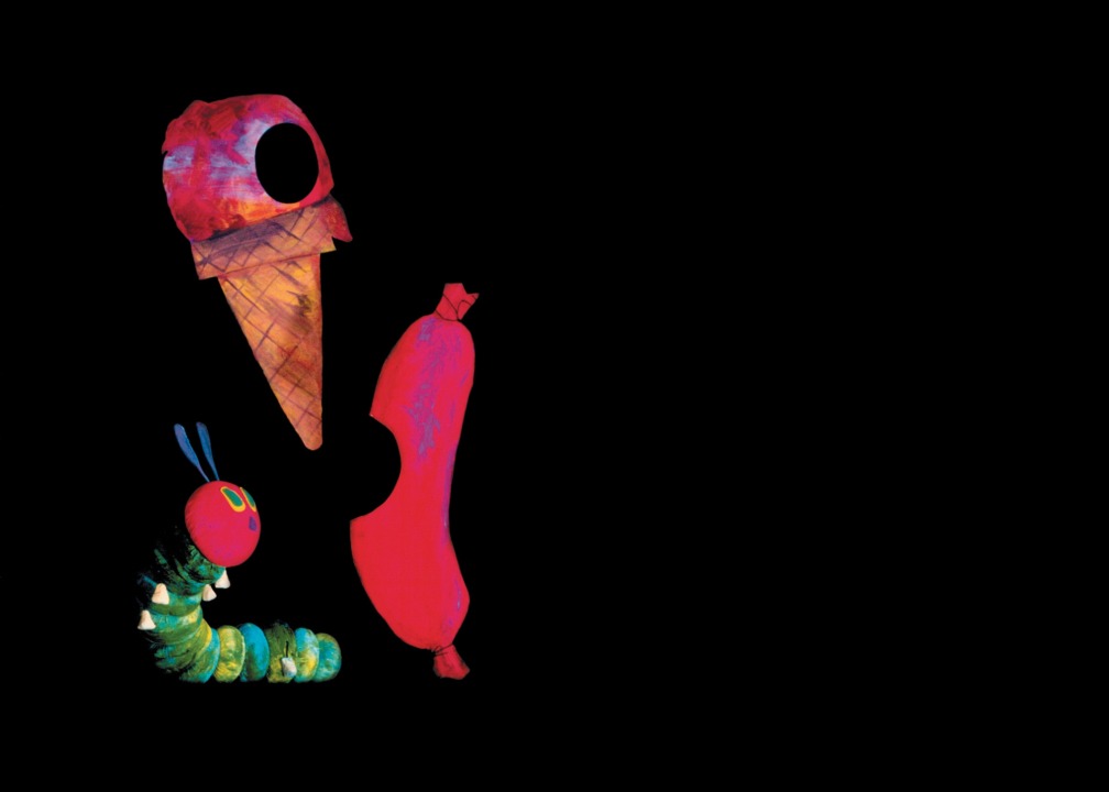 The very hungry caterpillar puppet looking at a giant ice cream cone and sausage with holes where he had "eaten" and a black background