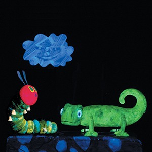 The very hungry caterpillar puppet looks up at a cloud prop with a green chameleon puppet next to him