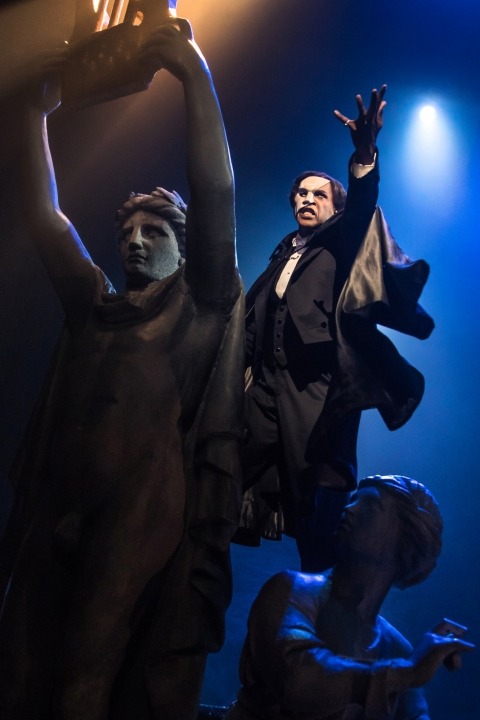 Phantom dressed in black with a white mask standing on two statues reaching out in front of a blue background.