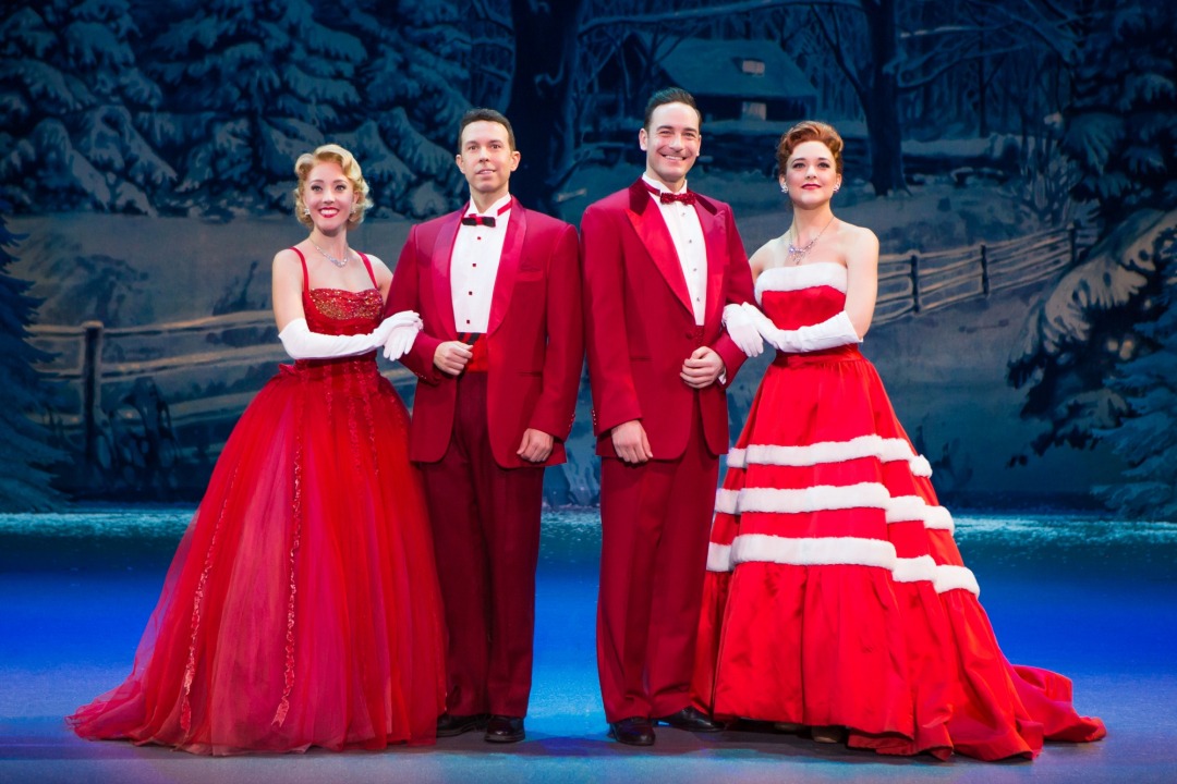 Two couples dressed in red standing arm in arm in front of a snowy cottage background