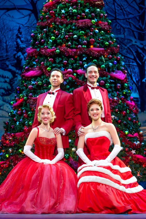 Two men standing behind two sitting women all dressed in red in front of a Christmas Tree