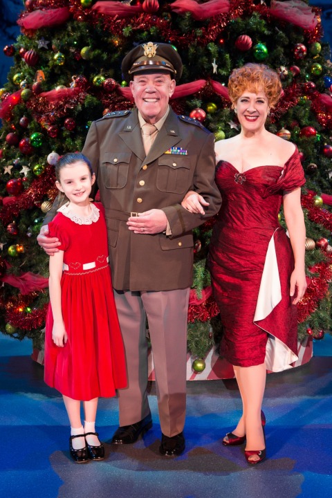 Man in uniform, woman in red dress, and young girl in red dress stand smiling in front of a Christmas Tree