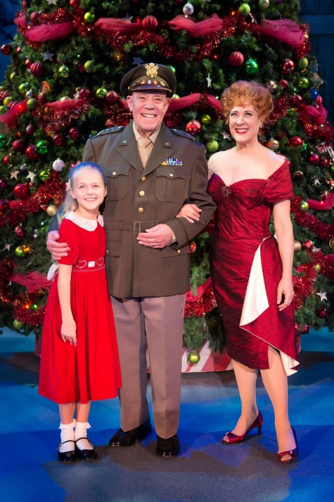 Man in uniform, woman in red dress, and young girl in red dress stand smiling in front of a Christmas Tree