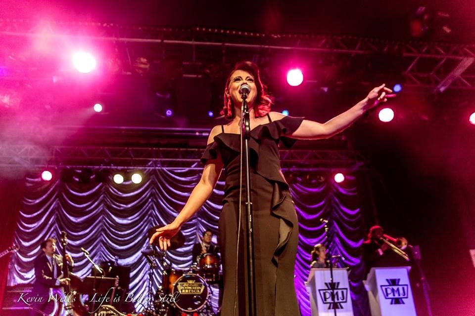 White woman, wearing a flowing black dress, singing into the microphone. Several pink lights in the background, as well as a purple fabric back-drop. Four other band members (trombone, violin, drums, and bass) are visible behind the singer.