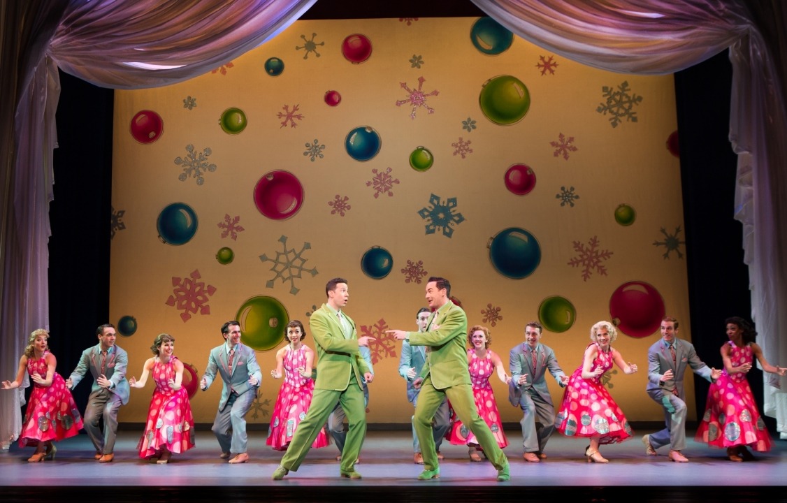 Two men in green shuts pointing at each other in front of a row of people in gray suits and red dresses dancing in front of a yellow background with ornaments and snowflakes on it