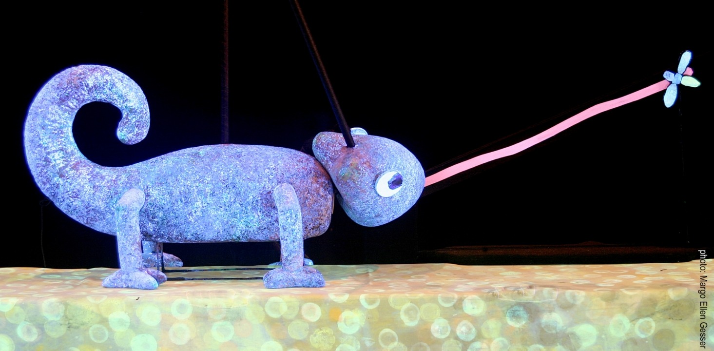 A purple chameleon puppet catching a fly with its tongue in front of a black background