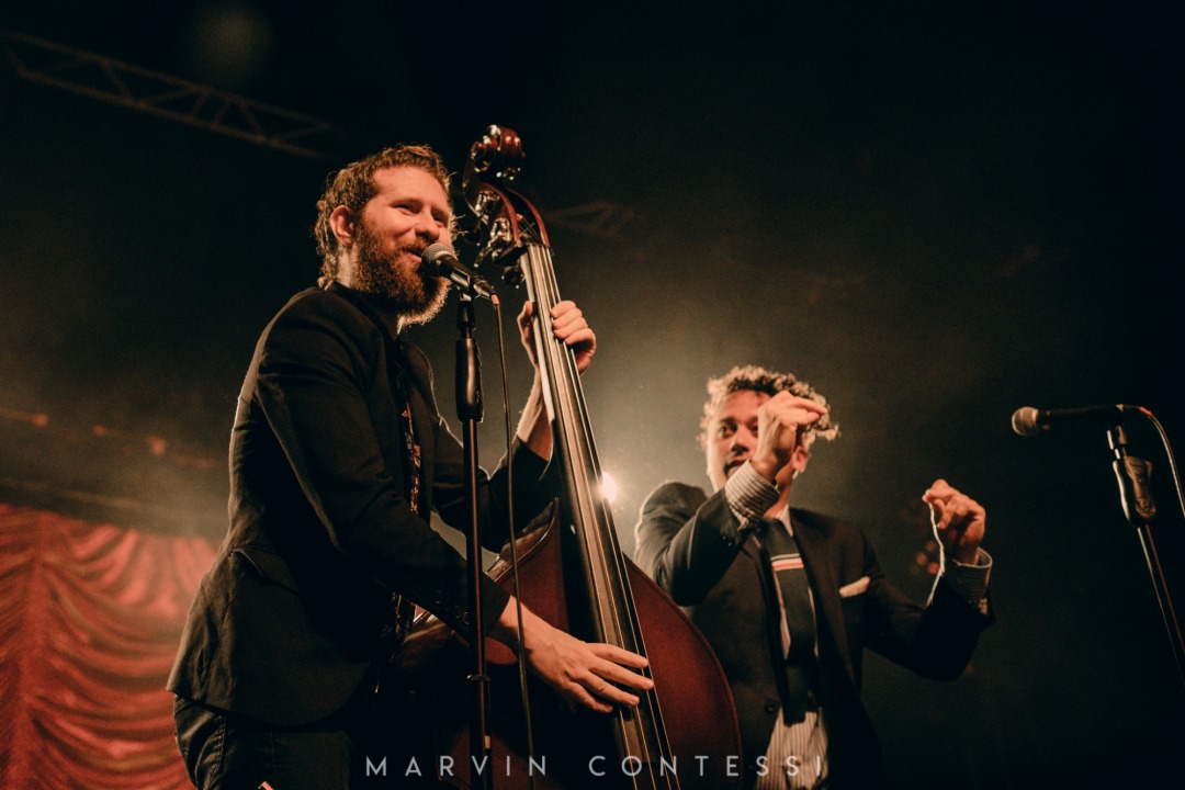 Two men wearing black suits, one with red hair and a beard and the other with curly brown hair. The red head is singing into the microphone and holding a stand-up bass. The man with brown hair is snapping his fingers.
