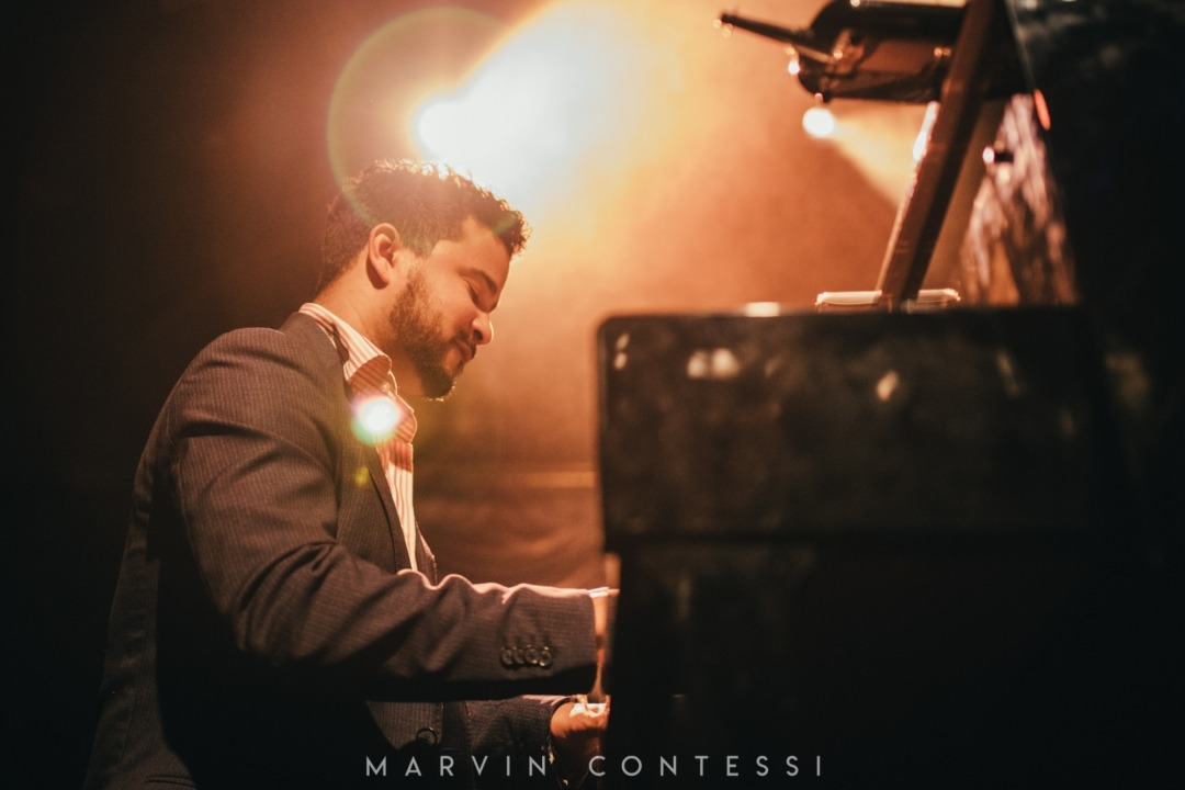 Side angle of man wearing a grey suit jacket with a red and white striped collared shirt, playing the piano. Bright yellow light in background.
