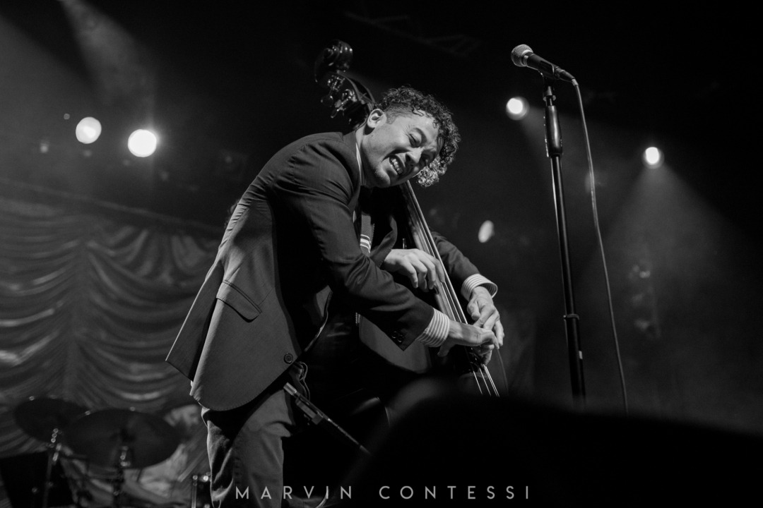 A black and white image of two individuals playing on the same bass instrument - with only one person's body visible (due to the angle of the photograph). Part of a drum set is visible in the left bottom corner of the image.