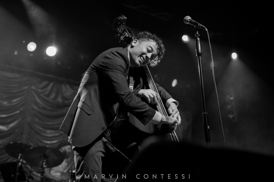 A black and white image of two individuals playing on the same bass instrument - with only one person's body visible (due to the angle of the photograph). Part of a drum set is visible in the left bottom corner of the image.