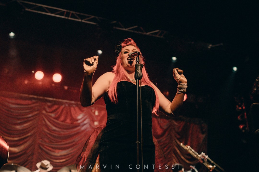 White women wearing a strapless black dress, has pink hair, and wearing many bead bracelets on her left hand, stands in the middle of the image singing into the microphone. Part of a drumset and trombone in the background of the image. Red fabric background.