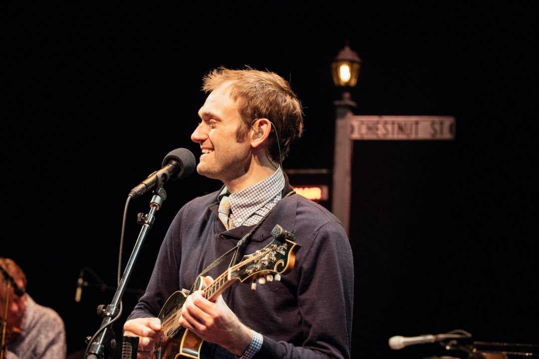Chris Thile sings and plays mandolin wearing a collared shirt and sweater.