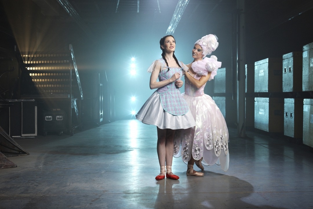 Woman in pink dress and pink hair with a woman in a blue dress and red shoes in a room with a blue light behind her